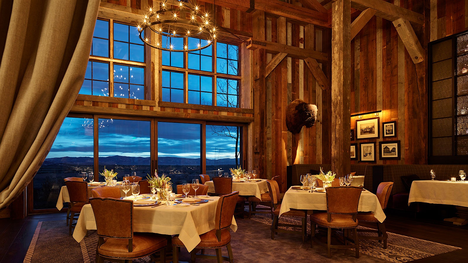 A dining room at Cheynne Club, with a bison trophy on the wall, wooden rafters and leatherbound chairs; large windows offer a view of mountains